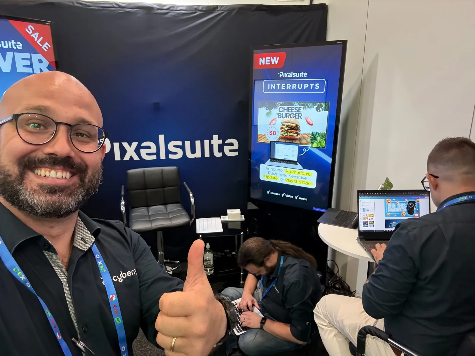 Julian Pennisi giving a thumbs up at the Cyberry PixelSuite stand at an industry expo, with the team at work behind him