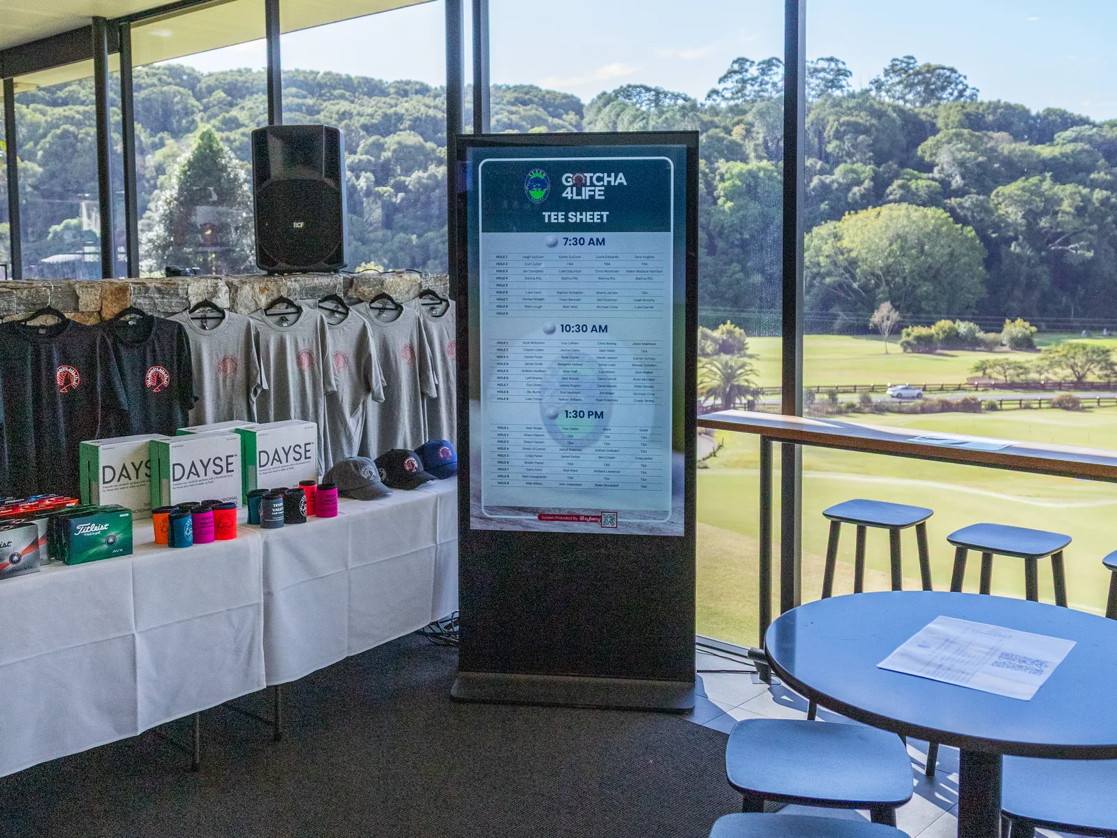 A freestanding PixelSuite display showing the tee sheet at a golf club, beside the event merchandise table