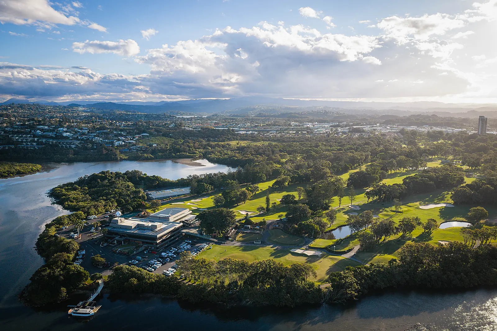 Late-afternoon aerial of Coolangatta & Tweed Heads Golf Club — the clubhouse on the bend of the Tweed River with the West Course fairways beyond and the hinterland ranges on the horizon