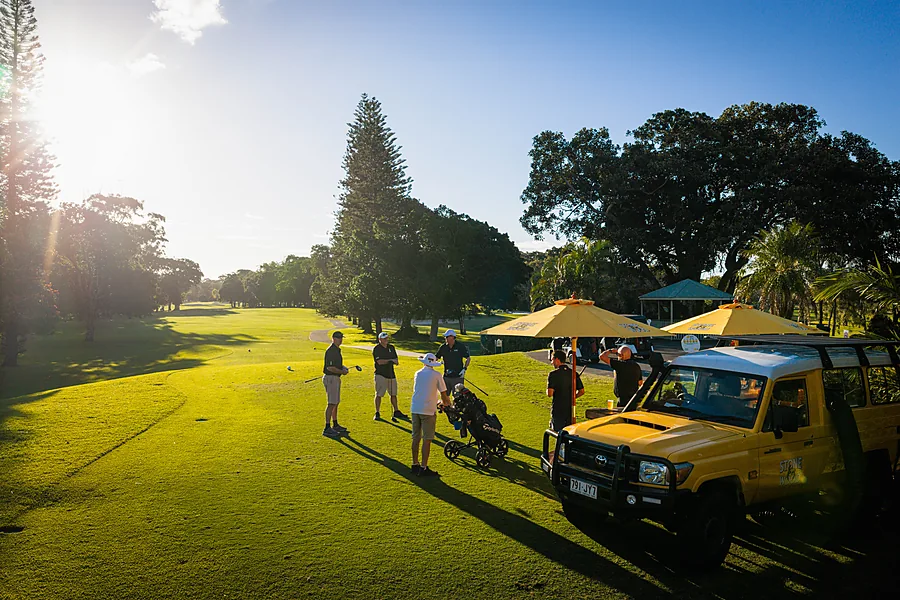 Backlit ground-level shot of players and the yellow drinks cart on the fairway as the sun drops behind the tree line