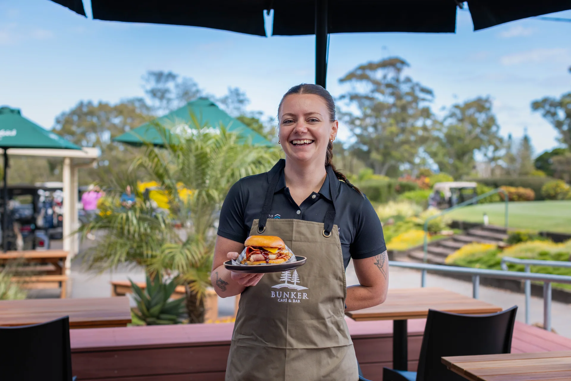 A smiling staff member at Bunker Café & Bar, Ballina Golf Club, holding out a burger on the terrace